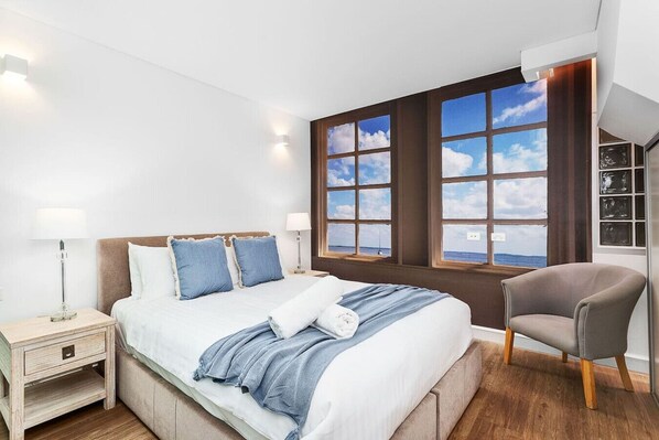 A modern bedroom with a large bed, two bedside tables, a chair, and a view of the sea through large windows. - Nest on Newcastle – T3 (Perth)