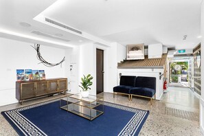 A modern lobby with a glass coffee table, a dark blue sofa, a wooden cabinet with magazines, and a potted plant. - Nest on Newcastle – T3 (Perth)