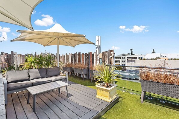 A rooftop patio with a wooden deck, a sofa, an umbrella, and potted plants.