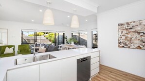 A modern kitchen with a central island, white cabinetry, and a dishwasher. There are pendant lights above the island and a large window providing a view of the outdoors. - Spacious & modern Dianella! (Dianella)