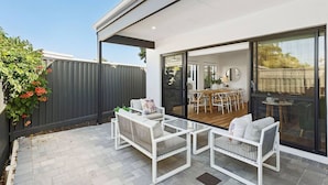 An outdoor patio with white furniture, a dining area with a wooden table and chairs, and a view through large glass doors. - Spacious & modern Dianella! (Dianella)