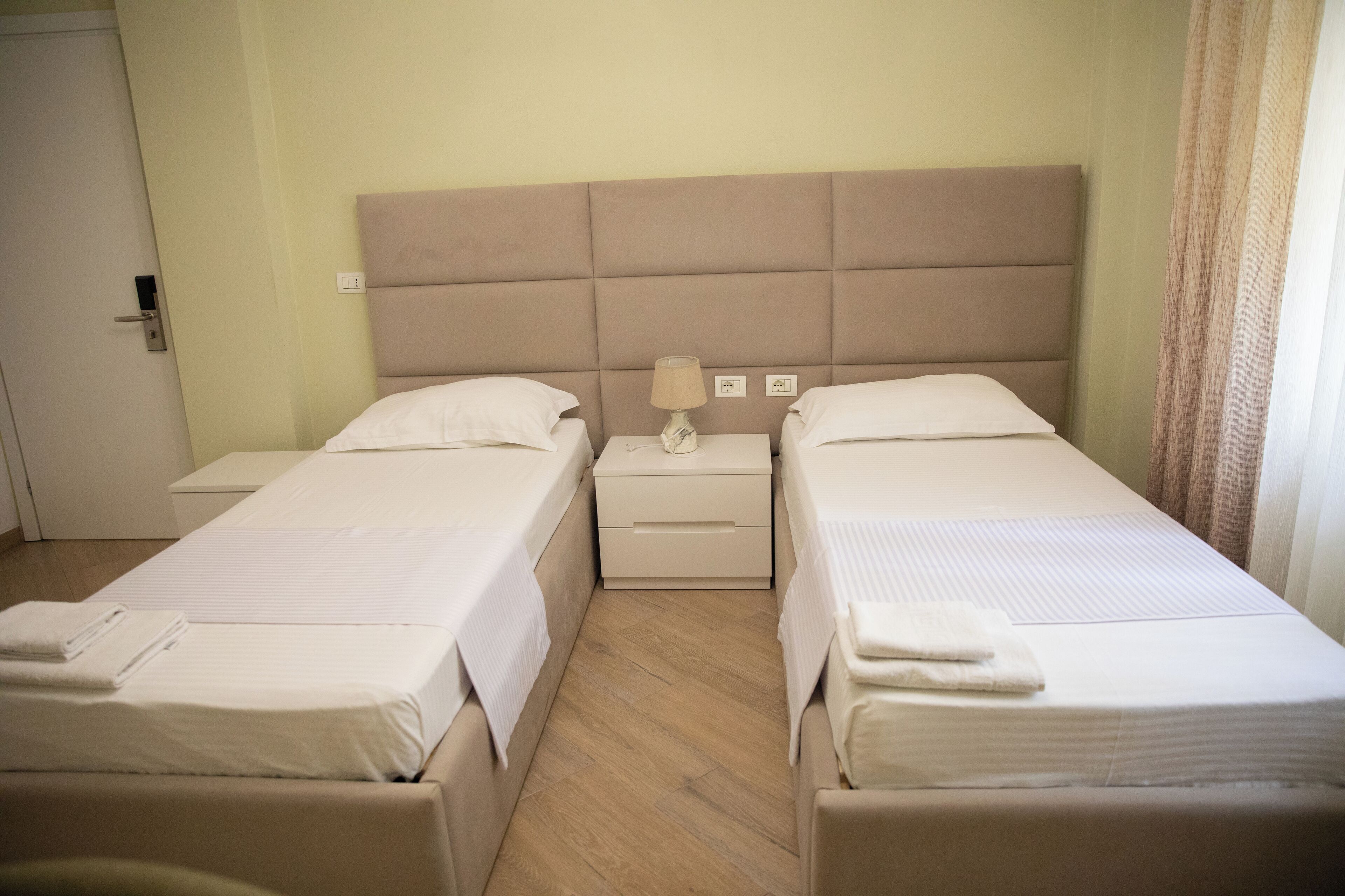 A hotel room with two single beds, a nightstand with a lamp, and a door.