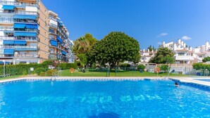 Een zwembad met helderblauw water, omringd door een hek, en een meerlaags appartementencomplex op de achtergrond.