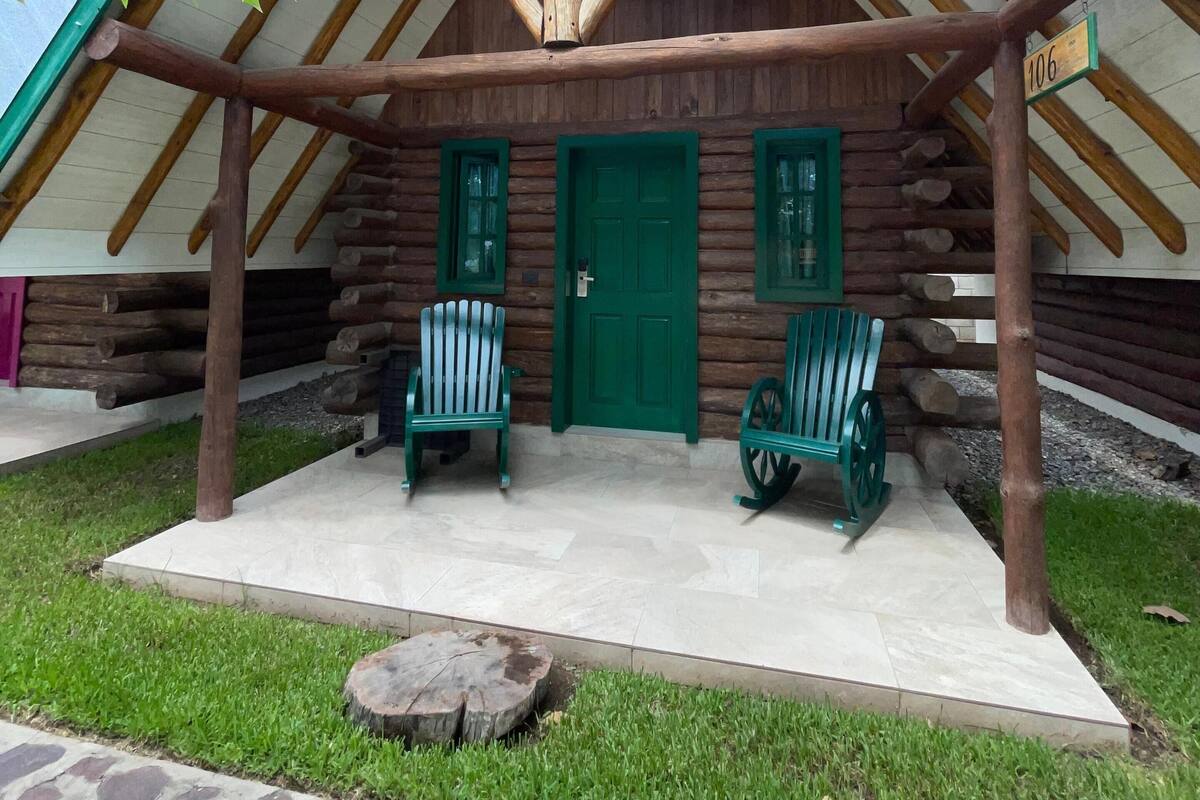Una cabaña de troncos con techo inclinado, dos ventanas con marco verde y una puerta verde. Dos sillas Adirondack verdes están afuera.