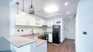 Una cocina moderna con armarios blancos, un refrigerador negro y un horno empotrado. Hay luces colgantes con diseño tejido, y el piso es de madera.