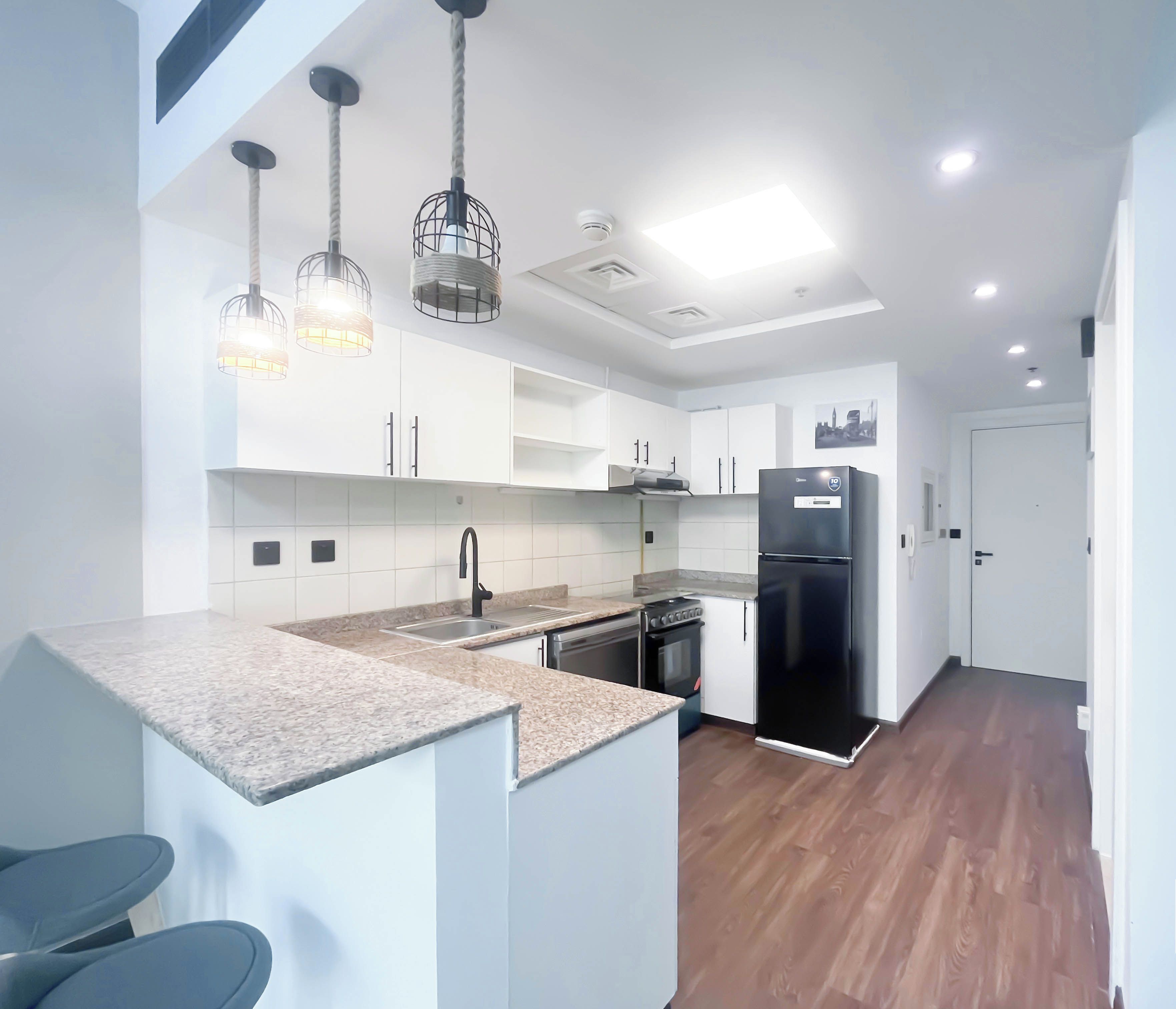 A modern kitchen with white cabinets, a black refrigerator, and a built-in oven. There are pendant lights with a woven design, and the floor is wooden.