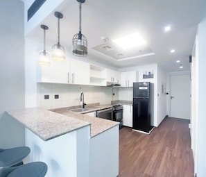 Een moderne keuken met witte kasten, een zwarte koelkast en een ingebouwde oven. Er hangen pendelverlichting met een geweven design en de vloer is van hout.