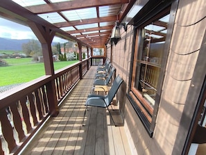 A covered outdoor seating area with wooden flooring, metal railings, and a view of a green landscape. - Vacation apartment with pony farm 🐴🐶🐓 (Edertal)