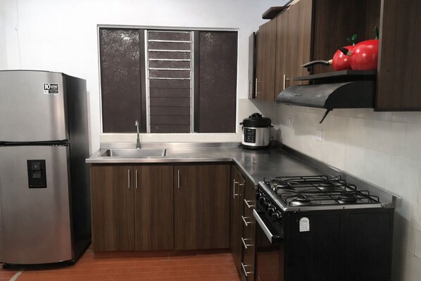Una cocina con refrigerador de acero inoxidable, estufa a gas, armarios de madera, ventana con persianas y una manzana roja en la repisa.
