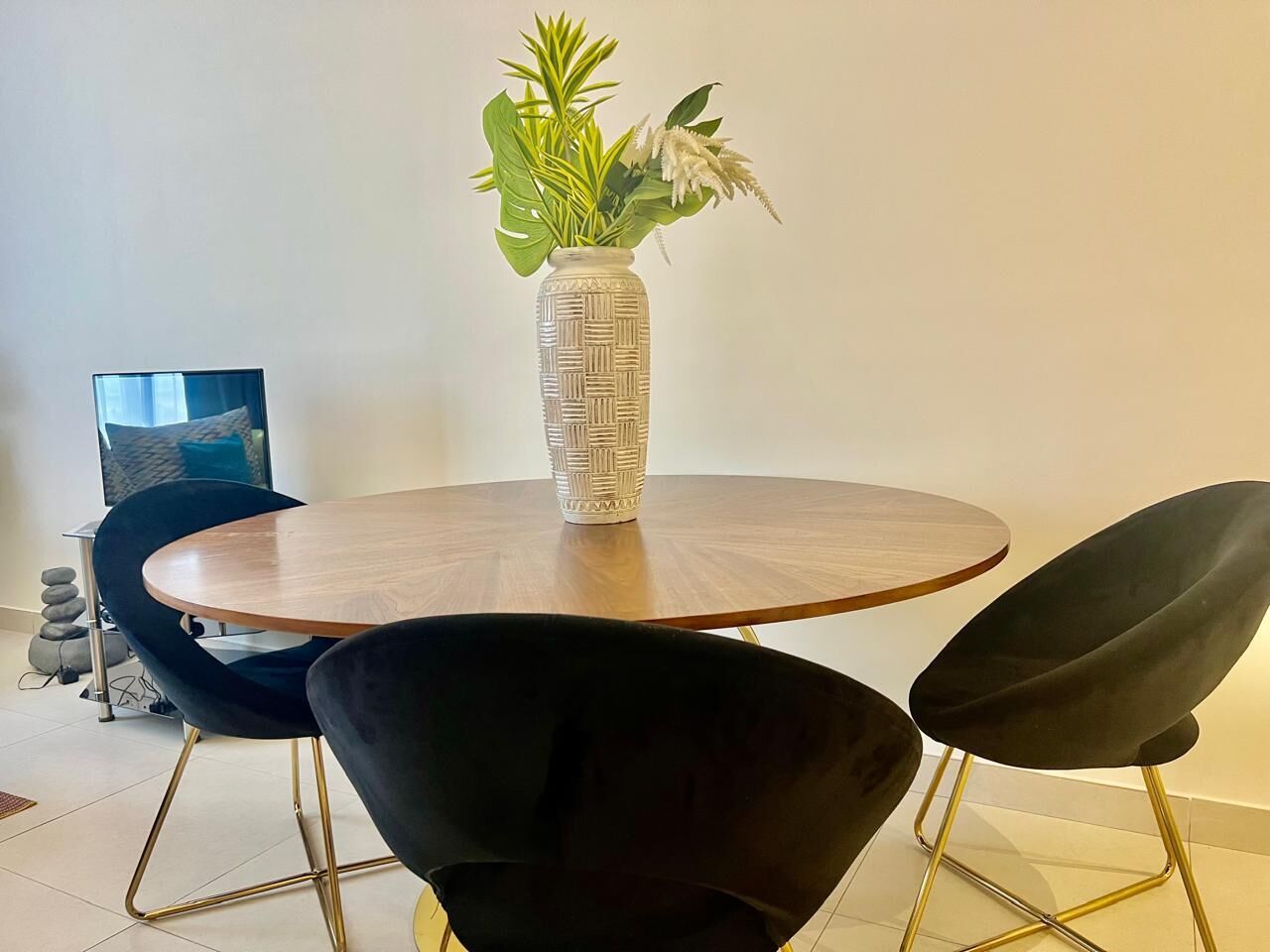 Una mesa redonda de madera con un florero tejido lleno de flores, dos sillas negras con patas doradas y una silla moderna con cojín negro.