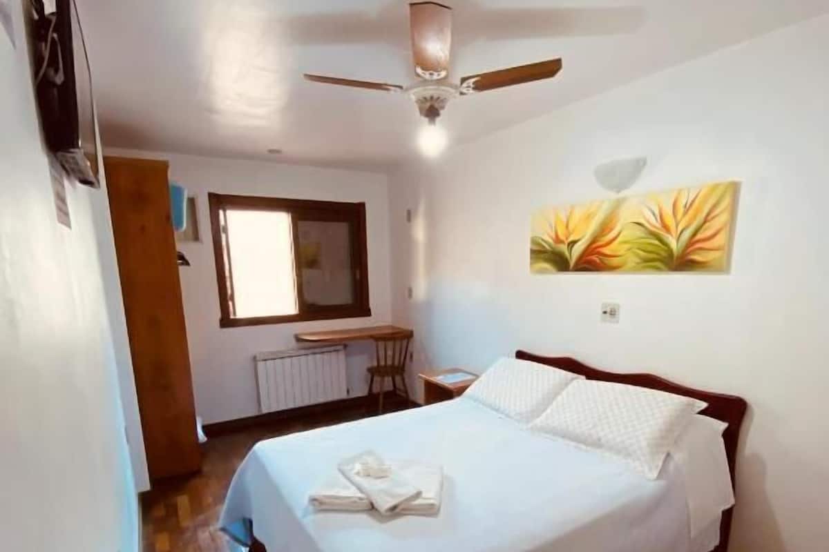 A bedroom with a bed, a ceiling fan, a window, and a painting on the wall.