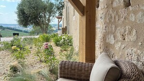Une chaise en osier avec un coussin, placée dans un espace extérieur abrité, avec un mur en pierre et une vue sur un jardin et un paysage lointain.