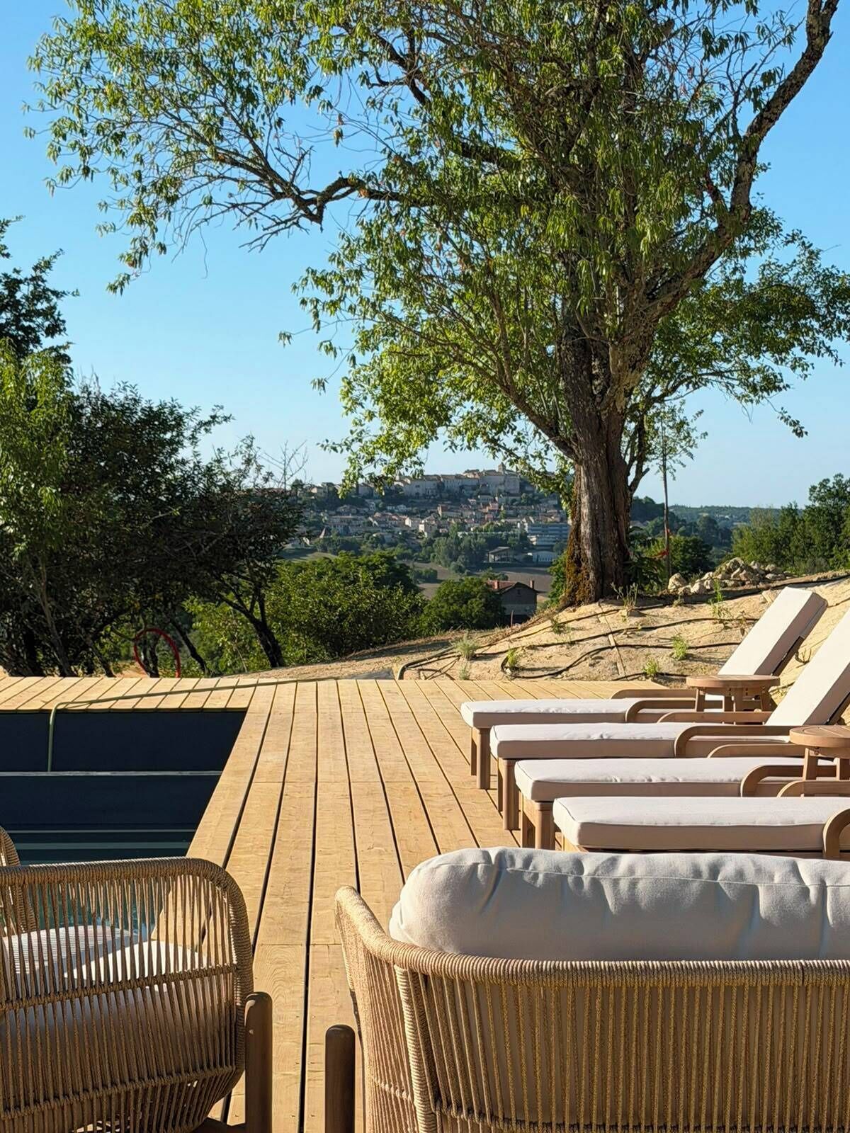Una terraza de madera con sillas de descanso y una silla de mimbre, con vista a un paisaje pintoresco de un pueblo y un árbol grande.