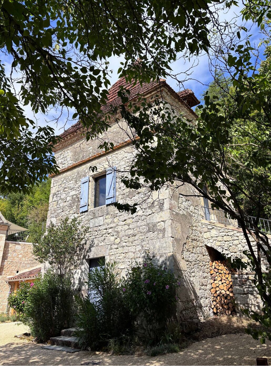 Un bâtiment en pierre avec un toit en tuiles, un petit porche et une pile de bois de chauffage appuyée contre le mur.