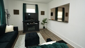 A living room with a dark-colored sofa, a black ottoman, a flat-screen TV, a dark entertainment unit, a potted plant, and a large mirror on the wall. - Comfortable 4BR Home Near Downtown Elyria (Elyria)