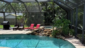 Une piscine agrémentée d’une cascade, entourée d’une terrasse couverte avec des chaises longues rouges et un barbecue.