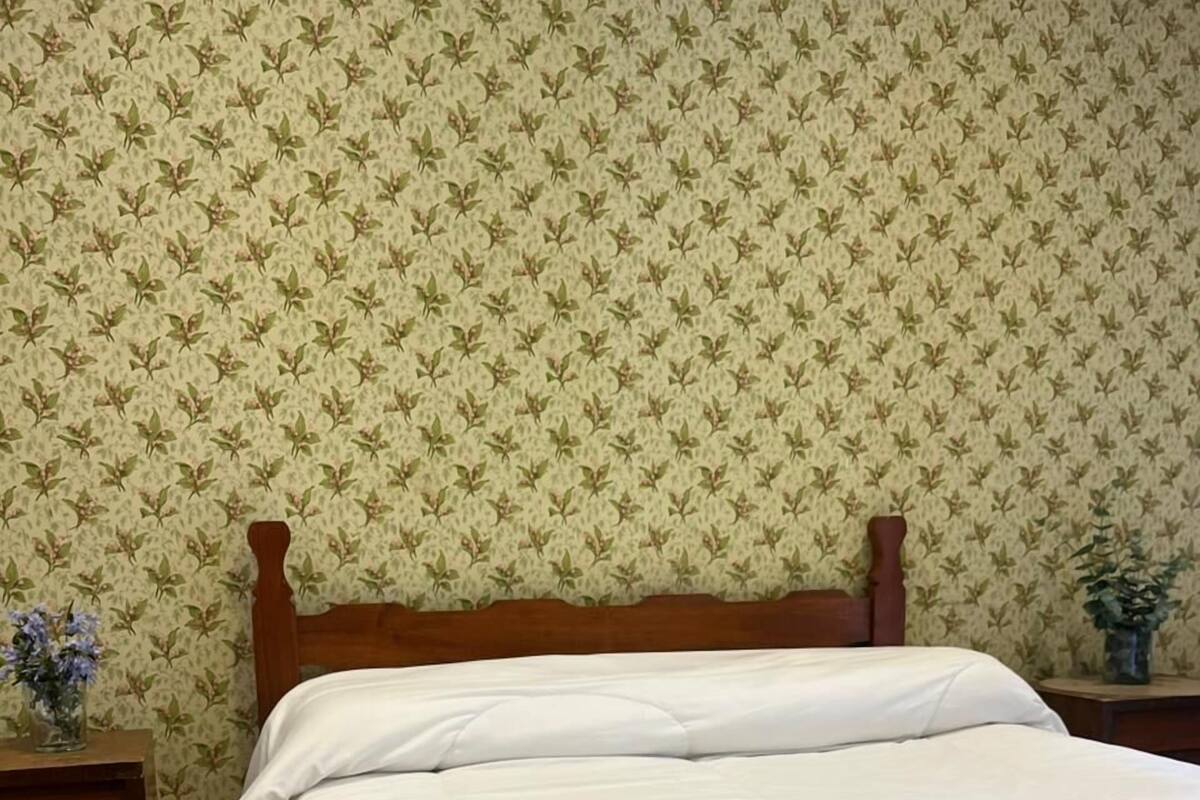 Quarto com cama de madeira, roupa de cama branca e papel de parede com padrão floral.