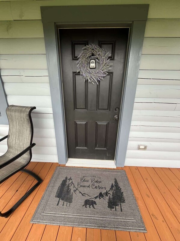 Une porte en bois sombre ornée d’une guirlande, un paillasson gris avec des silhouettes d’animaux, et une chaise grise sur la gauche.
