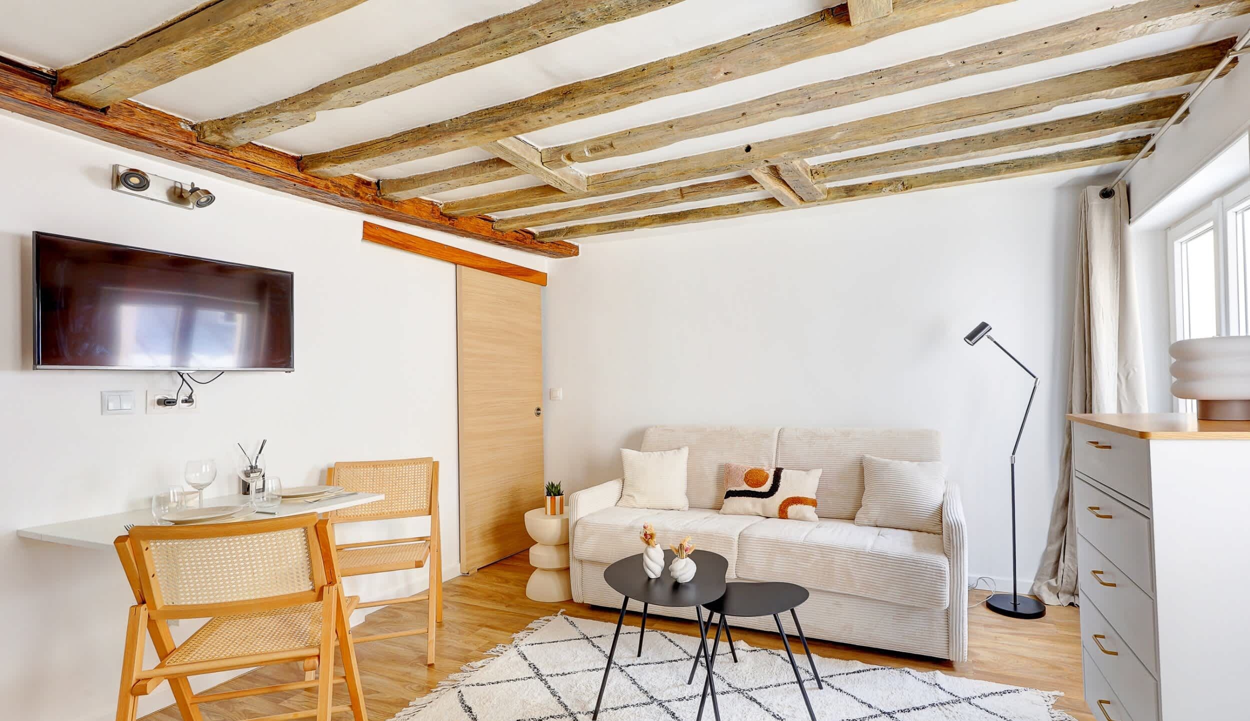 A modern living room with a wooden ceiling, a flat-screen TV, a white sofa, a dining table with chairs, and a small coffee table.