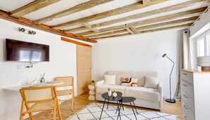 A modern living room with a wooden ceiling, a flat-screen TV, a white sofa, a dining table with chairs, and a small coffee table. - BM - Studio refait à neuf en plein coeur du Marais (Paris)