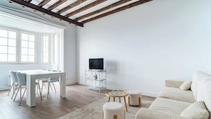 A spacious living room with a white sofa, a dining table with chairs, a TV on a stand, and a wooden ceiling.