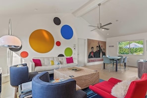 Una sala de estar moderna con un sofá blanco, sillas azules y rojas, una mesa de centro y una zona de comedor con mesa y sillas.