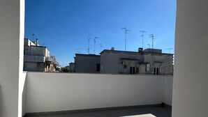 A rooftop terrace with a clear blue sky, tiled flooring, and buildings in the background.