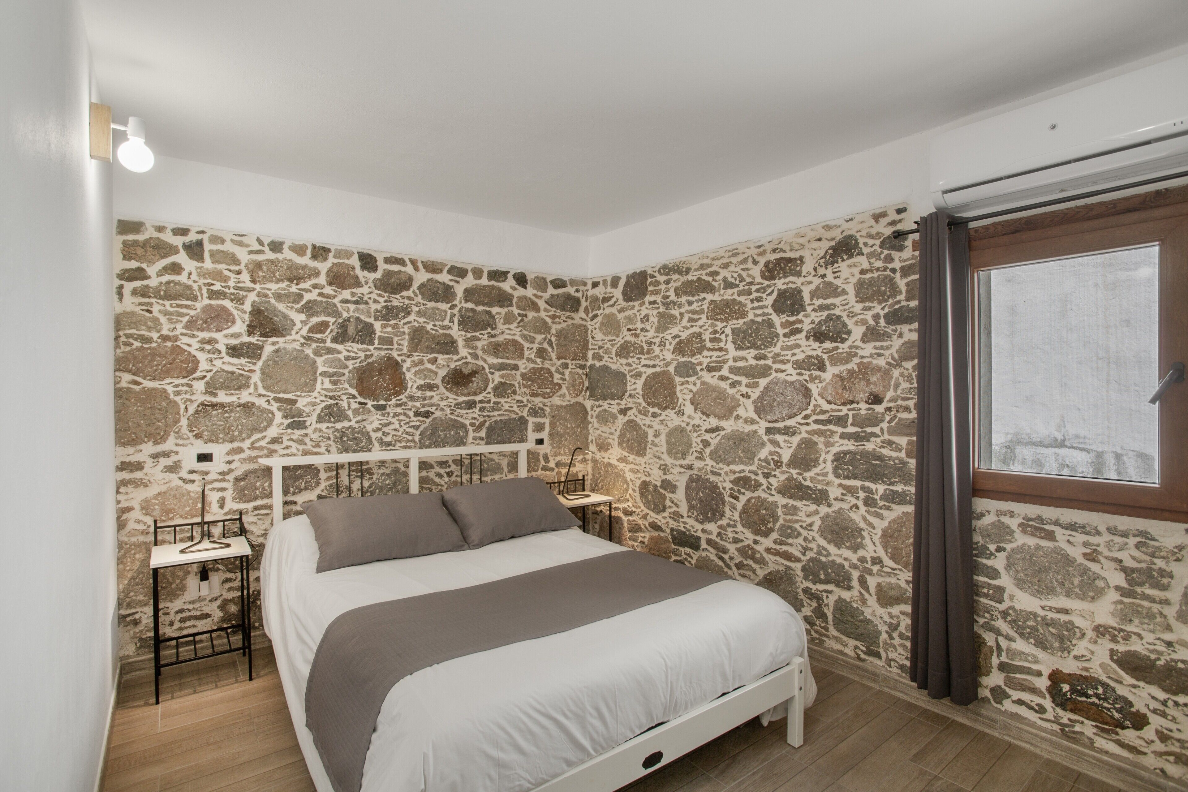 Un dormitorio con pared de piedra, cama con cabecera blanca, mesita de noche y ventana con cortinas.