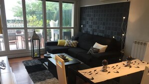 Ein modernes Wohnzimmer mit einem schwarzen Sofa, gemusterten Kissen, einem Esstisch mit Unterlage und Blick ins Freie durch große Fenster.