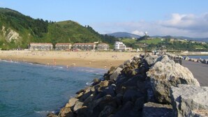 Ein Küstenstrand mit einem felsigen Wellenbrecher, einem sandigen Ufer und Gebäuden im Hintergrund.