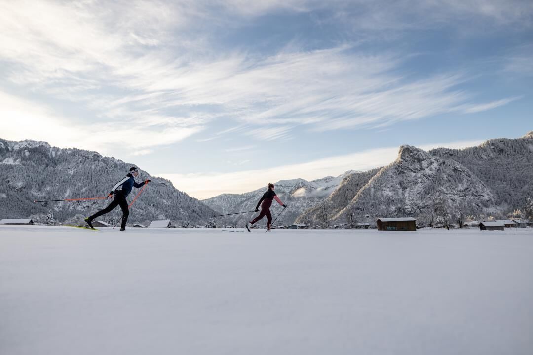Duas pessoas praticam esqui cross-country em uma paisagem nevada, com montanhas ao fundo.