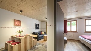A room with a wooden ceiling, a sofa, a bed, a small table with a vase, and a window with curtains.