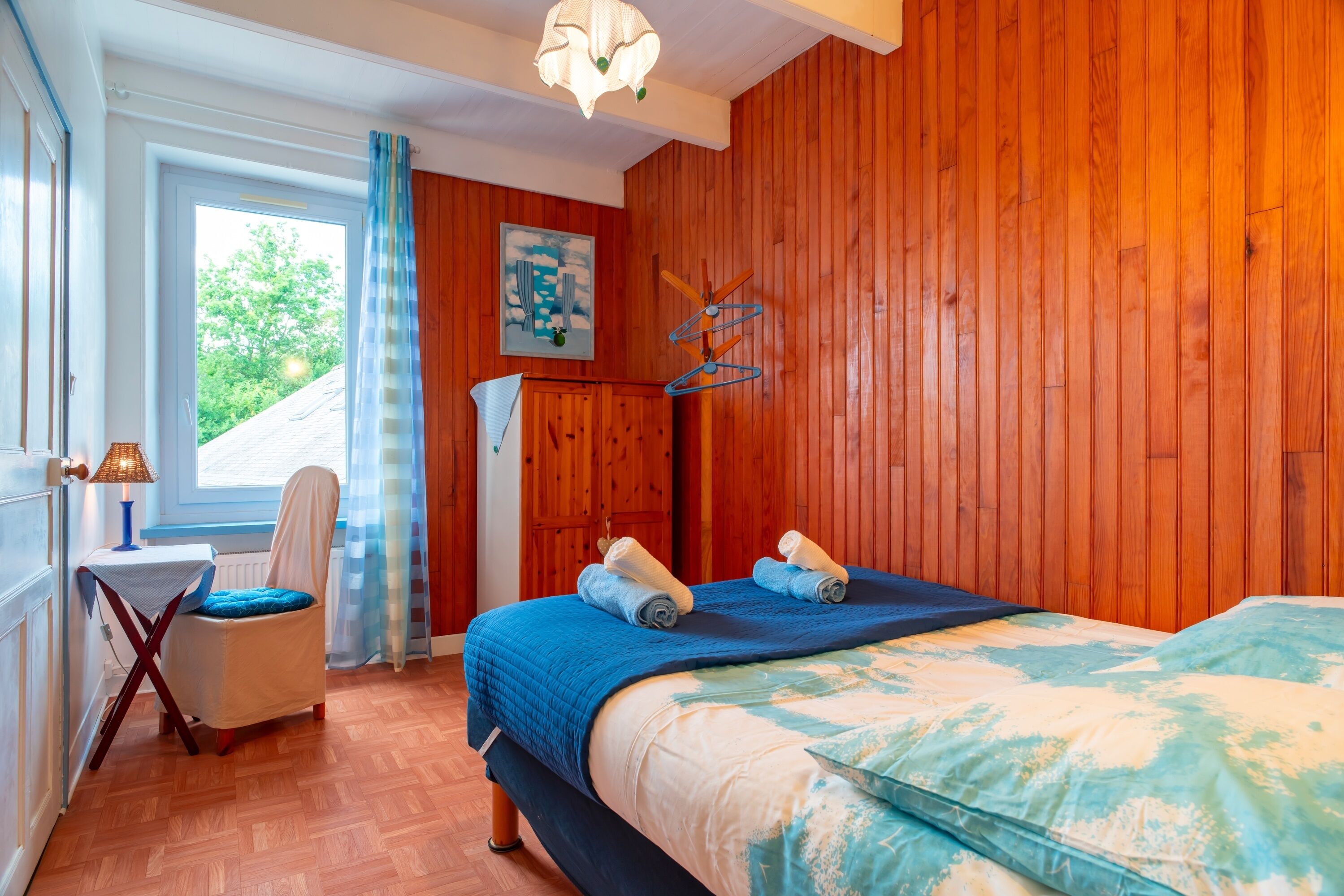 A room with wooden paneling, a bed with blue bedding, a chair with a blue cushion, a table with a lamp, and a window with a view of greenery.