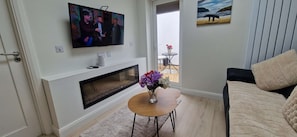 A living room with a TV mounted on the wall, a fireplace, a small round table with a vase of flowers, and a couch.