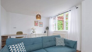 A living room with a blue sofa, two patterned pillows, a small table, a lamp, and a window with curtains.
