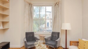Een hoekkamer met twee grijze fauteuils, een klein rond tafeltje, een vloerlamp, een bank met een geel kussen en een boekenplank.
