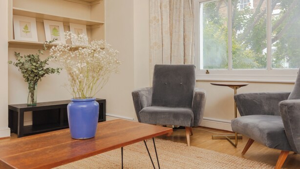 Een woonkamer met een houten salontafel, twee grijze fauteuils, een blauwe vaas met witte bloemen en een boekenkast met ingelijste foto’s.