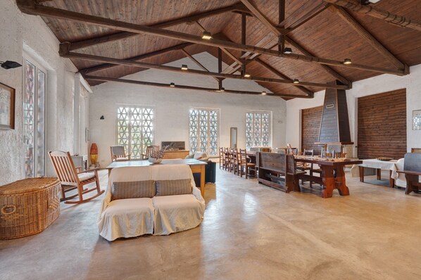 A spacious room with a high wooden ceiling, large windows with lattice designs, a central dining table, and a cozy seating area with a sofa and chairs.
