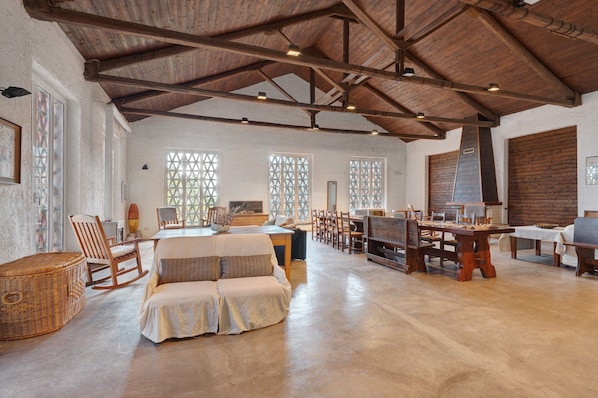A spacious room with a high wooden ceiling, large windows with lattice designs, a central dining table, and a cozy seating area with a sofa and chairs.