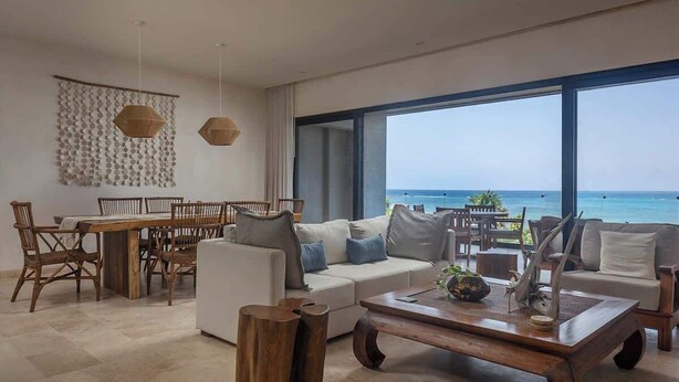 Uma sala de estar moderna com uma grande janela vista para o oceano, um espaço de jantar com móveis de madeira e uma área de estar acolhedora com almofadas.
