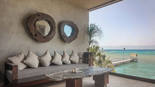 A modern room with a large window overlooking turquoise waters, a wooden sofa with cushions, and a unique table with a natural pattern. - Beachfront Luxury + Rooftop Oasis Tankah Bay (Tulum)