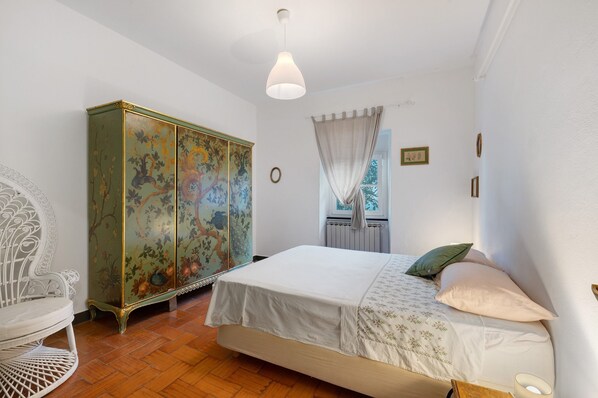 Un dormitorio con cama, un aparador decorativo, una silla y una ventana con cortinas.