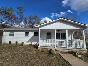 A two-story house with a porch, a small garden, and a clear blue sky. - Charming Mountain Retreat with fire pit, fenced yard, scenic views, and privacy. (Mineral Bluff)