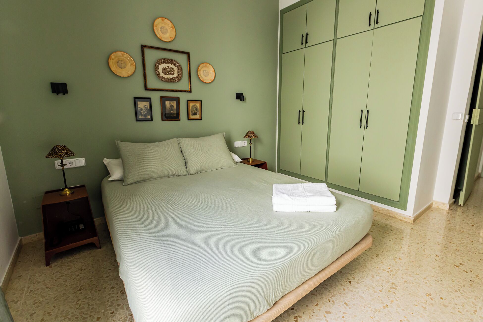 A bedroom with a bed, green wardrobe, framed pictures, and two bedside tables with lamps.