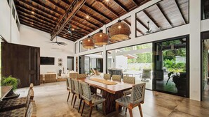 A spacious dining area with a large wooden table and chairs, a ceiling fan, and large windows offering a view of the outdoors.