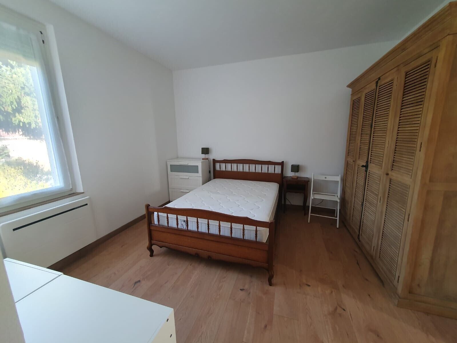 Een slaapkamer met een houten bed, een nachtkastje, een bureau, een kledingkast en een raam.
