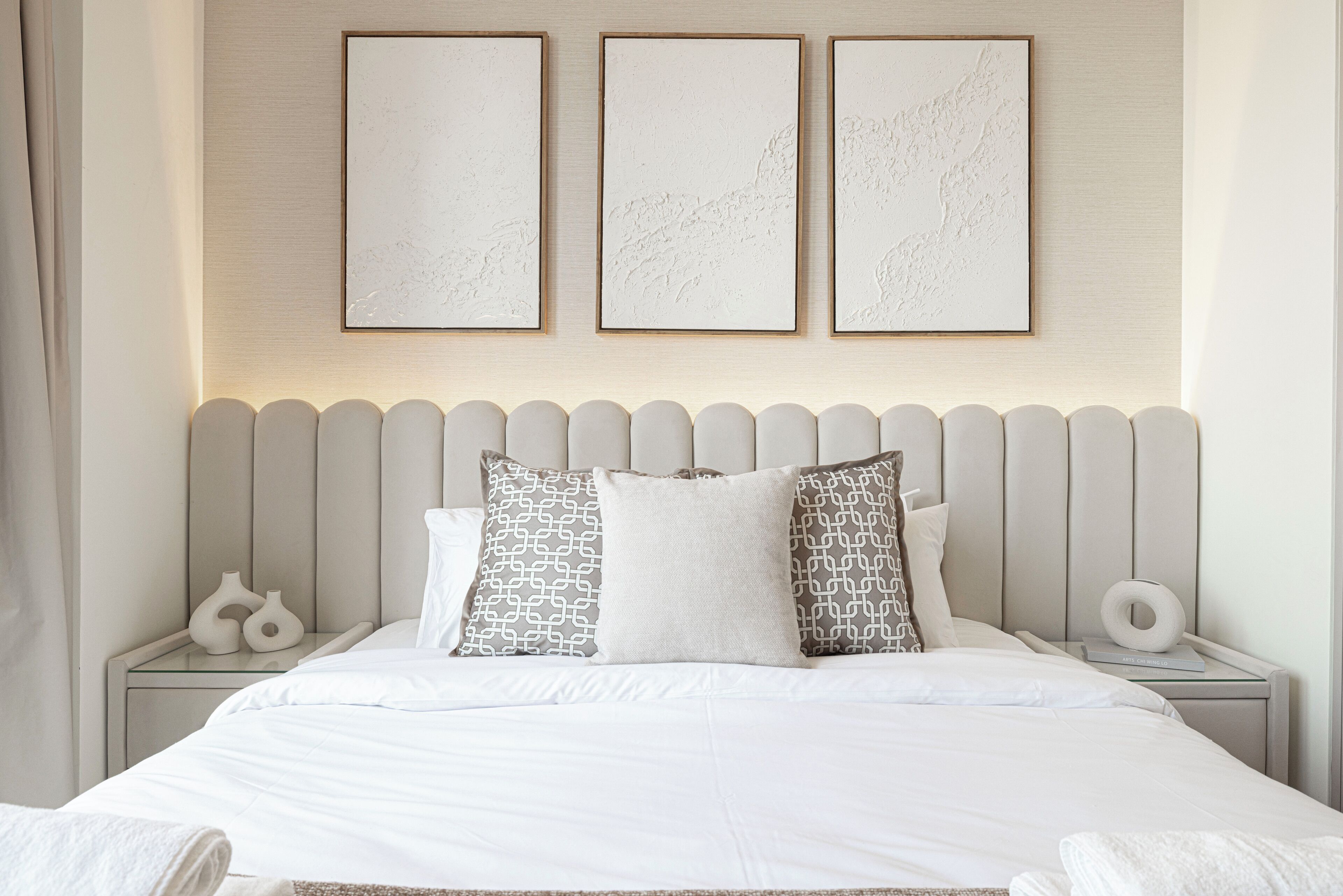 A neatly made bed with white linens and patterned pillows, a headboard with vertical slats, and three framed artworks on the wall above.