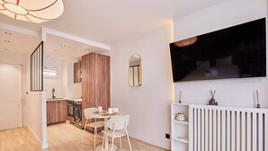 A modern kitchen with wooden cabinets, a dining area with two white chairs, a large flat-screen TV mounted on the wall, and a white radiator. - THE Parisian SUITE / Center of Paris (Paris)