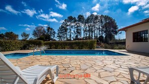 A swimming pool with stone tiles, white lounge chairs, and a view of tall trees under a clear sky.