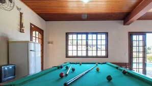 Una mesa de billar con bolas, un televisor y ventanas con vistas a la vegetación.
